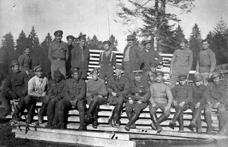 Black and white photograph. Members of the Canadian Forestry Corps sit for a group photo on top of building materials. Their uniforms are of differing quality; both black and white men are part of the group.